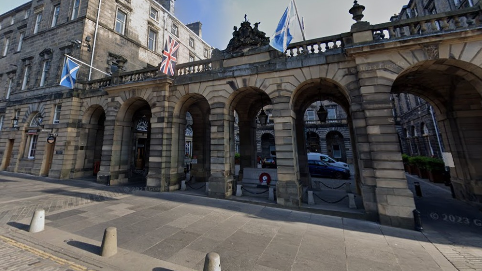 The front of Edinburgh City Chambers has stone pillars with flags hanging from it. Through the archways leads to a courtyard and the entrance to the City Chambers.