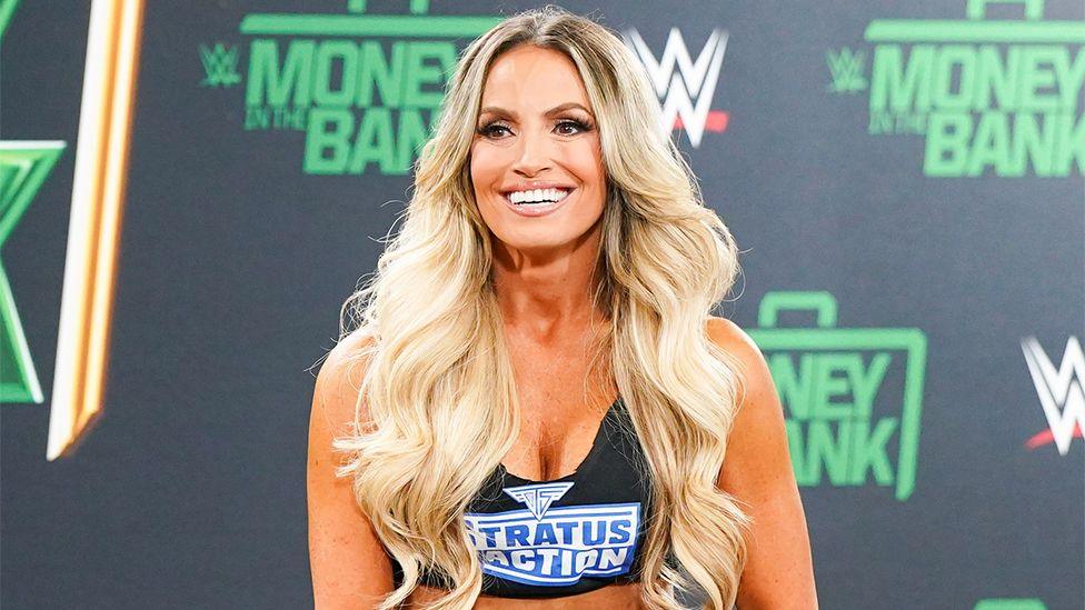 Trish Stratus, a woman, on stage at a WWE event for Money In the Bank event, wearing a black top which says "Stratus" in blue font. Behind her is a wallpaper of black with green font which reads Money in the Bank.