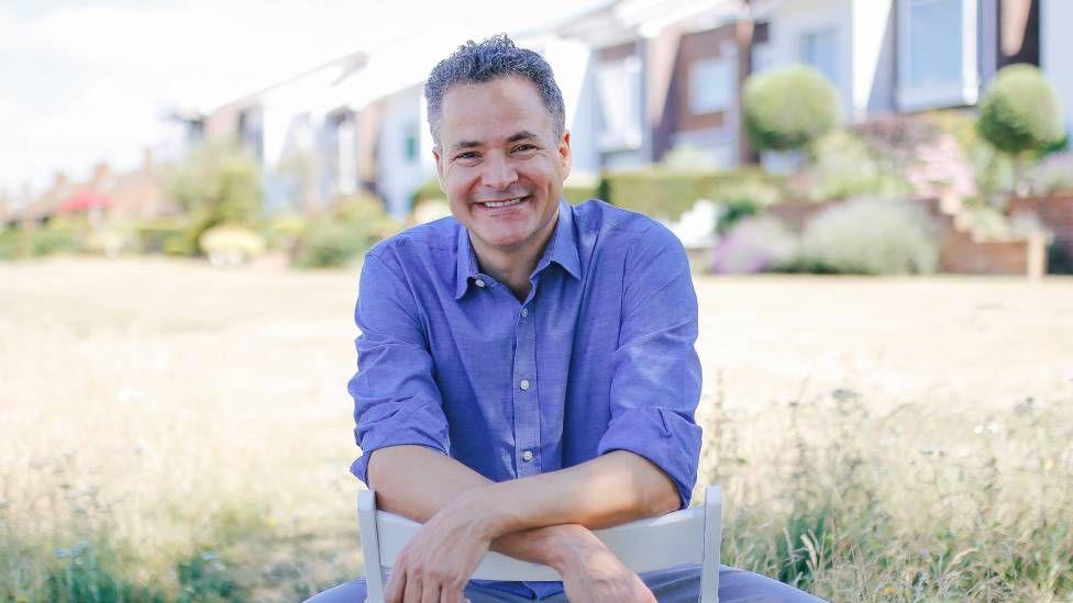 A smiling man with grey hear wearing a blue shirt sitting on a chair in a sunny garden