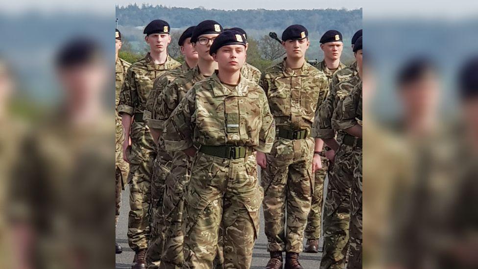 Gunner Beck wearing her camouflage military uniform and black cap. She is standing among her fellow soldiers with her arms clasped behind her back and a stern facial expression.