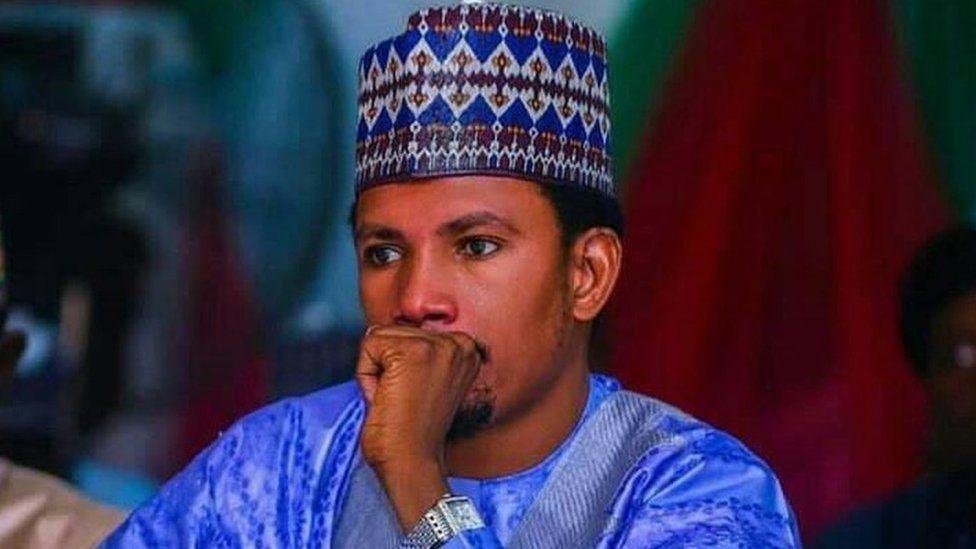 Elisha Abbo, sitting with his hand over his mouth, wearing a traditional blue cap and gown