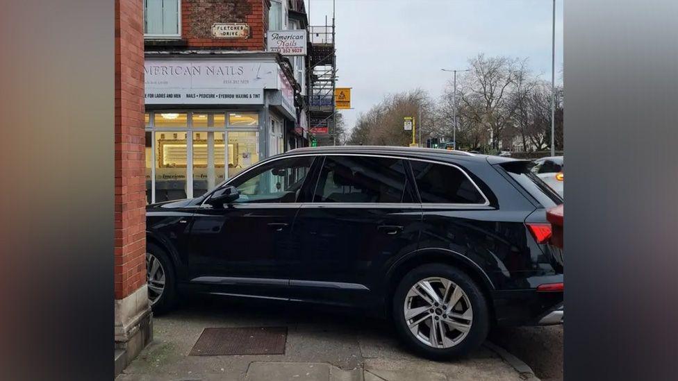 A black car parked on the corner of a street blocking access for pedestrians on the pavement