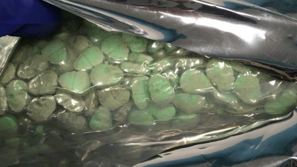 A clear bag contains lots of green pills in a skull shape. They are sealed in a foil bag