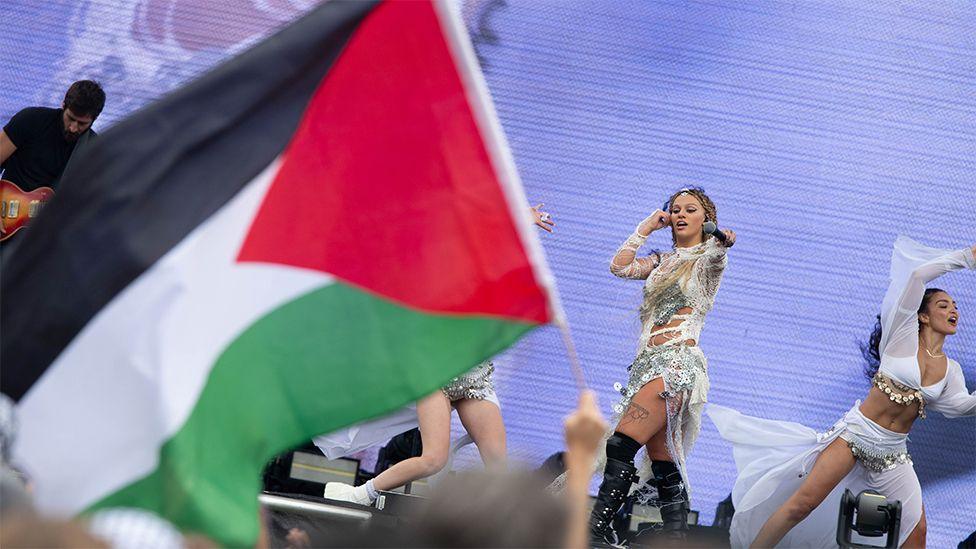 Elyanna performing on stage in San Francisco. She is wearing a white and silver glittery outfit, with her right hand going towards her right ear and her left hand is holding a black microphone outstretched towards the crowd. In the crowd, a fan is holding the flag of Palestine, which is coloured red, black, white and green.