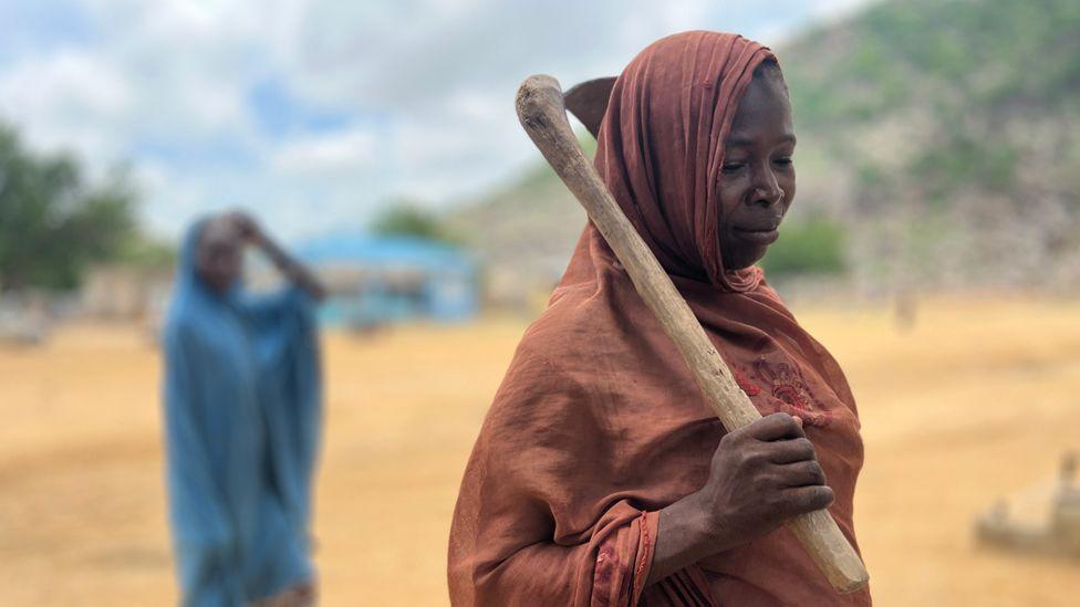 A woman in a long brown headscarf wrap holding a digging tool over her shoulder. Behind her to the left and out of focus is a woman in a blue head scarf wrap in Gwoza, Nigeria - July 2025.