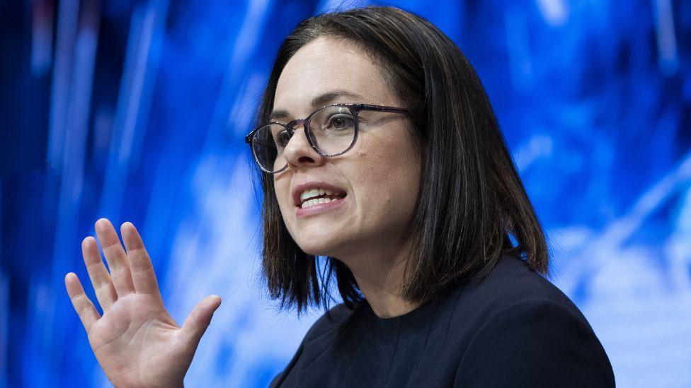 Kate Forbes, with dark hair and glasses and wearing a black top, speaks with one hand raised in front of a blue background in a close-up shot 