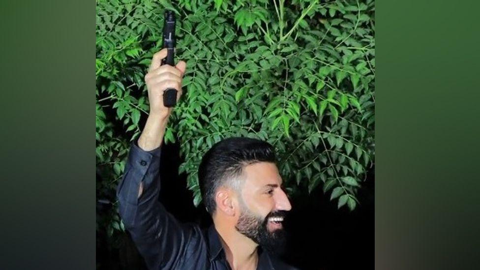 Amanj Hasan Zada, wearing a dark shirt and standing in front of a green leafy tree, holds a gun in the air while smiling.