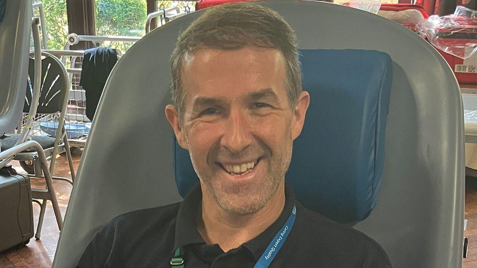 Stephen Kuypers sat in a grey hospital chair donating blood. He has short hair and stubble and is smiling
