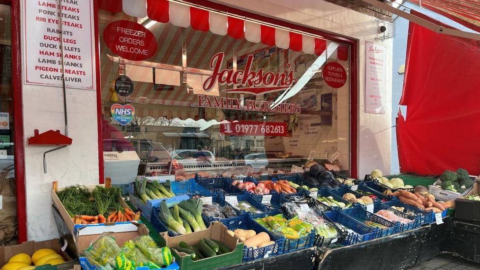 A red and white shop front which says "Jackson's family butchers".
There's baskets of vegetables laid out in baskets in front of the window including leeks, carrots, sweet potato, courgettes and broccoli.