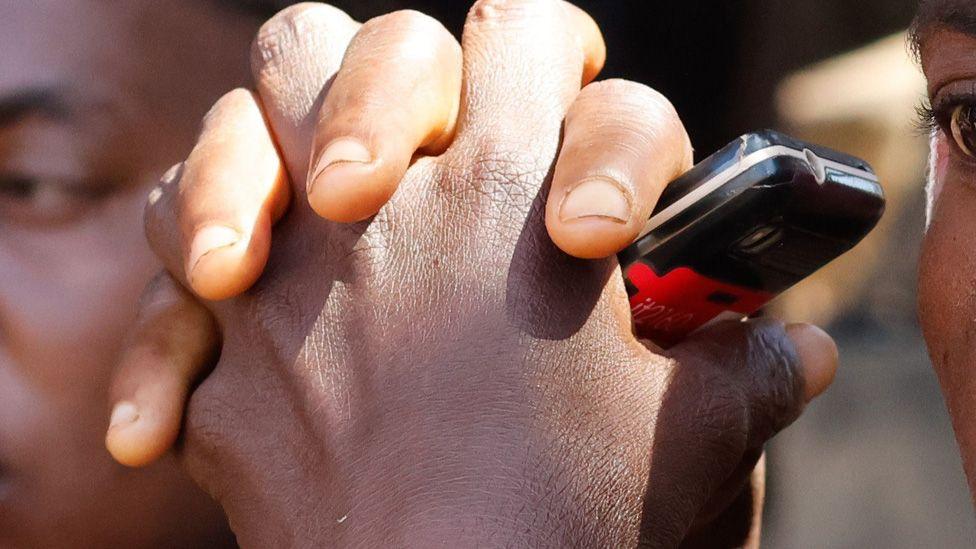 A cropped image showing clasped hands of a parent holding a mobile phone as they wait outside St Mary's Catholic School in Papiri, Niger State - 24 November 2025.