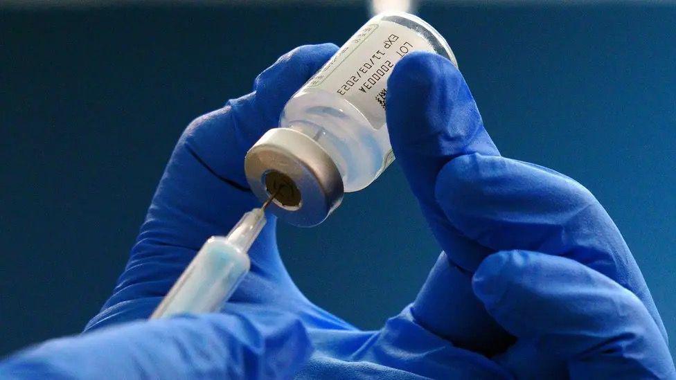 A syringe being pushed into a vial being held by a pair of hands wearing blue surgical gloves.