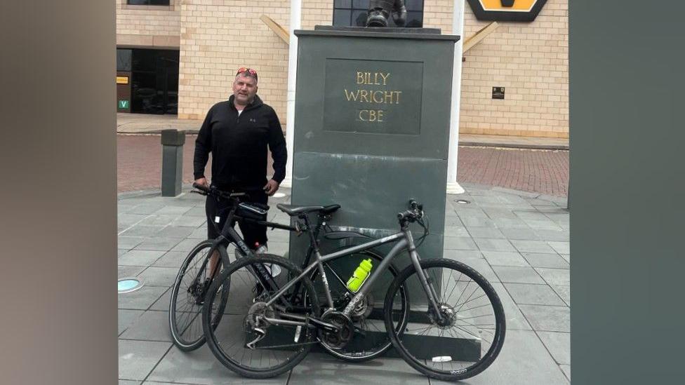 Ade Doughty is wearing a black top and black shorts. He is standing next to two bicycles. The bikes are leaning on a statue of Billy Wright CBE, which is outside the Wolverhampton Wanderers football stadium, which is visible in the background