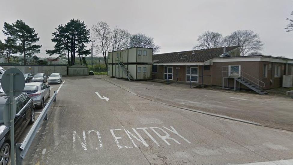 A Google streetview image of a laboratory building, garages and temporary buildings in Falmer. There is painted road signs reading no signal, a carpark to the left and a building and portakabins to the right of the image