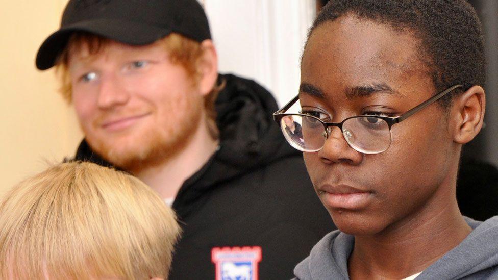 Ed Sheeran indoors wearing a black cap and black Ipswich Town jacket standing behind two boys