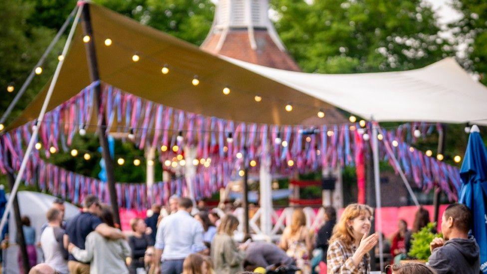 A tent with pink and blue streamers and tassels which has been put up in Chapelfield Gardens. There are fairy lights on the tent and crowds of people.