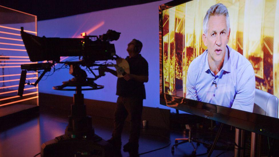 Gary Lineker seen presenting Match of the Day in the show's studio, next to a cameraman operating a large studio TV camera