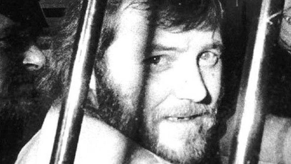 Andrew walker looking at the camera through bars. He has a beard. It is a black and white photograph.