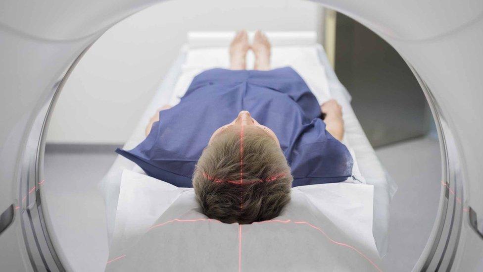 A patient in a navy blue hospital gown lies down inside a body scanning machine. A red laser is being used to create a cross in the middle of their head using two lines.