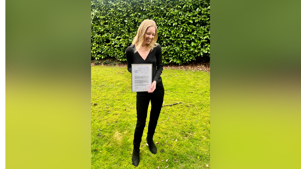 A tall, slim blond woman, Dr Emily Stacey, holds a framed copy of the letter that was replied to by Jimmy Carter. She wears a black top and trousers and is laughing, looking down away from the camera. She is standing outside on bright green grass with a hedge a few metres behind her. 