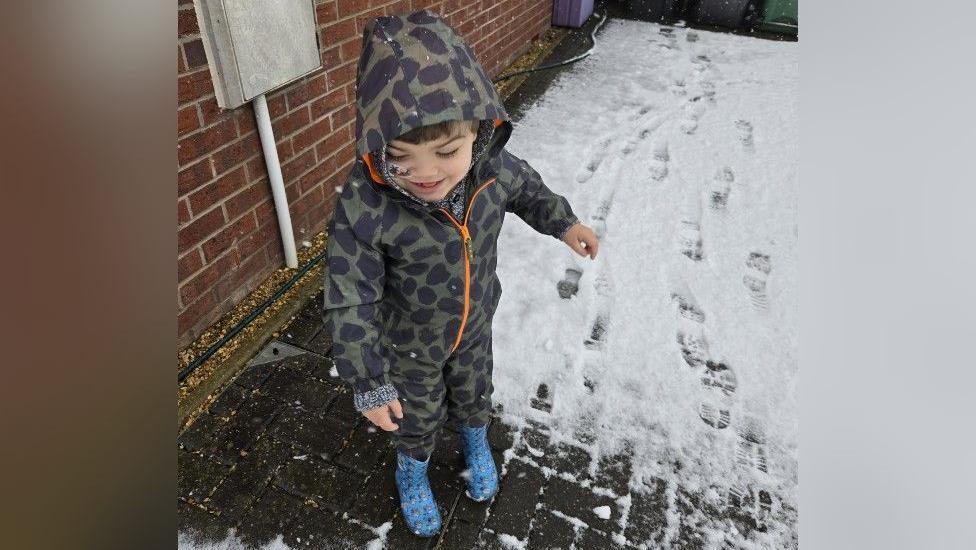 Gunner is wearing an all-in-one that is dark green with black spots. His hood is up and he is smiling. He is wearing blue wellington boots and is standing next to some snow on the ground