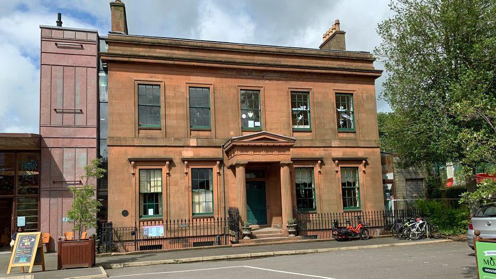 A Victorian mansion building in Dumfries made out of local sandstone with a few bicycles parked outside and a tree to one side of it