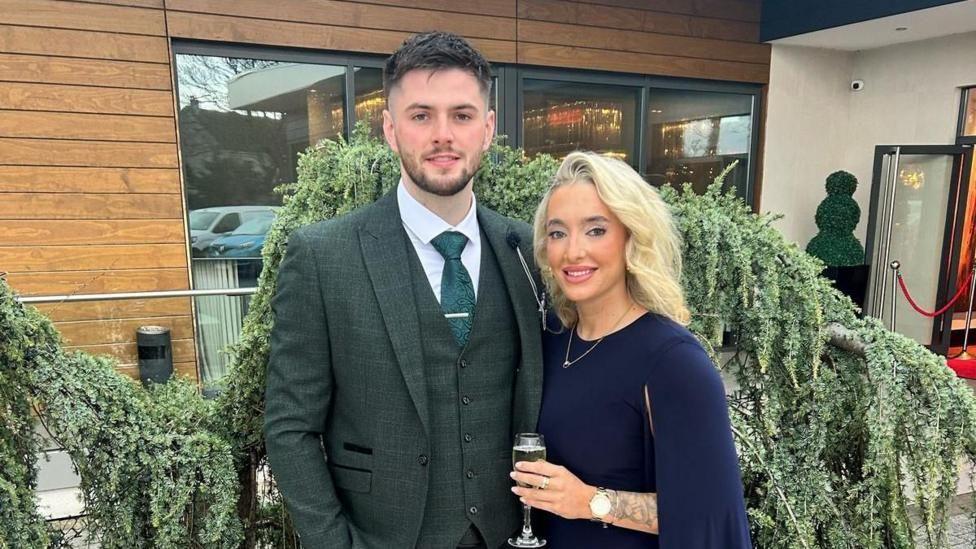 Man and a woman stand side by side. The woman, on the right, wears a navy dress with a gold necklace. She has blonde hair and make up on. She holds a glass of champagne in her left hand which has a gold watch on it. The man has a green tweed suit with green waistcoat and green tie. He has a white shirt and a tie bar on. He has short brown hair and a short brown beard. Behind them is a wooden building with large glass windows. A green bush sits between the couple and the building