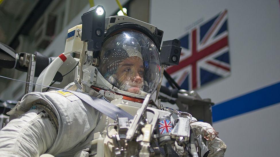 Close up picture of Rosemary Coogan in her full space suit, with her helmet on looking straight ahead just before she begins her dive. A light is attached to the helmet on the right hand side. A small Union Jack is visible on the front of her spacesuit and there is a large Union Jack flag that is partially visible on the wall over her left shoulder.