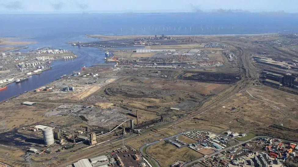An aerial view of the Teesworks site. The mainly undeveloped industrial land sits at the mouth of the River Tees as it enters the North Sea. Buildings are dotted around the land. There are dozens of wind turbines just off the coast.