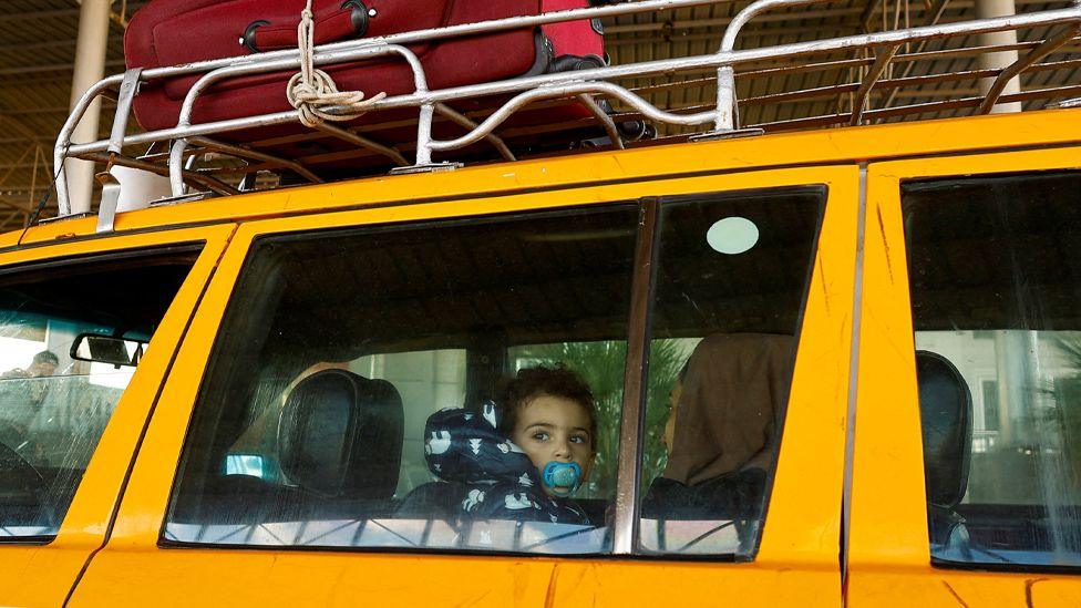 A Palestinian child, with a dummy in its mouth, looks out the window of a yellow car at the Rafah crossing in Egypt. The child is on the lap of a woman, whose face is turned away from the camera. Just visible on top of the car is a metal roof rack, with a red suitcase tied to it.