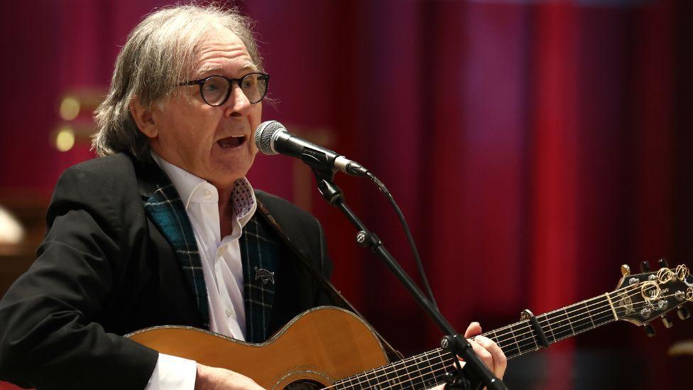 Singer Dougie MacLean, with grey hair, a dark suit and a guitar in hand, sings into a microphone while seated in front of a red background 