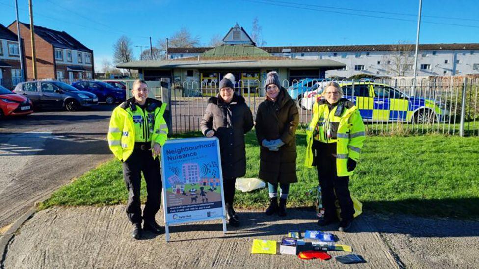 Two female police officers in high-vis uniforms stand either side of two council workers in dark coats and hats. In front of them is a sign saying "Neighbourhood Nuisance". In the background are houses and a police car.