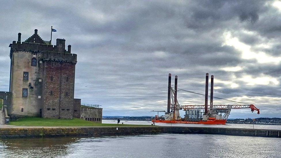 Legs of an oil platform sailing up a river on a boat, with an old castle in the foreground.