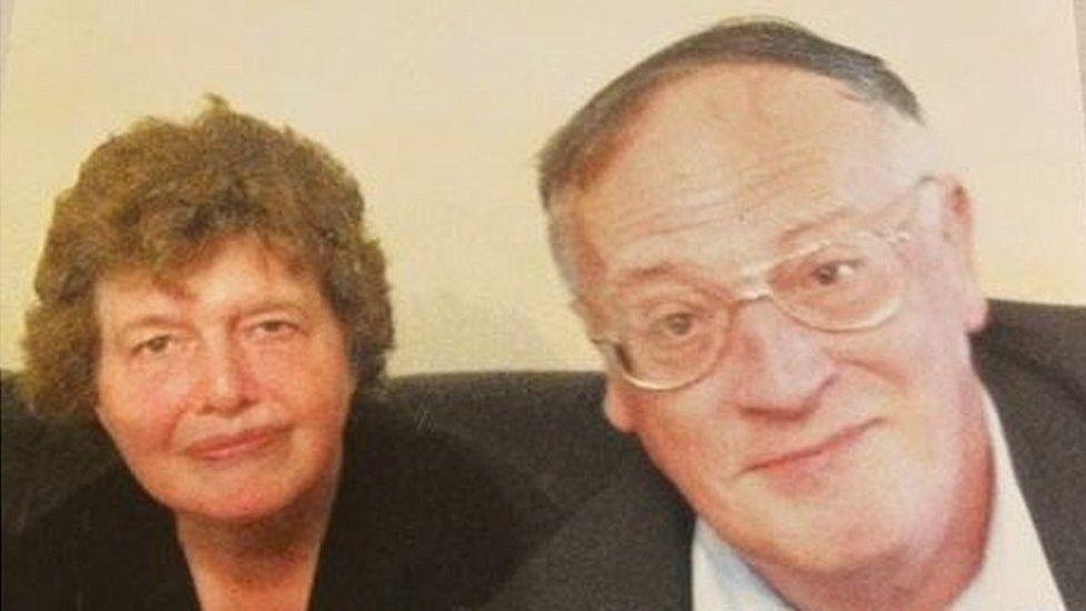 Lois and John McCullough. They are sat on a sofa and smiling at the camera.