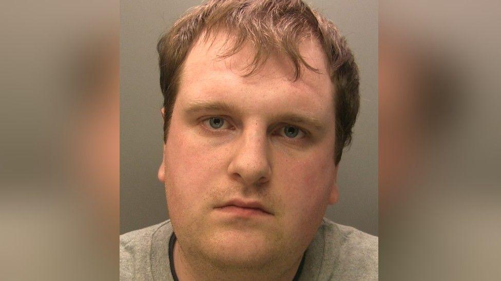 Police custody image of Jonsen Lamb. He has brown hair and is wearing a grey jumper. He is looking into the camera.