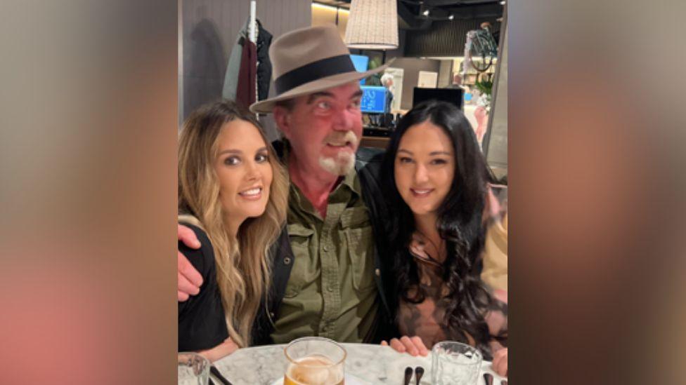 A man in a wide brimmed hat sitting in between two women in a pub