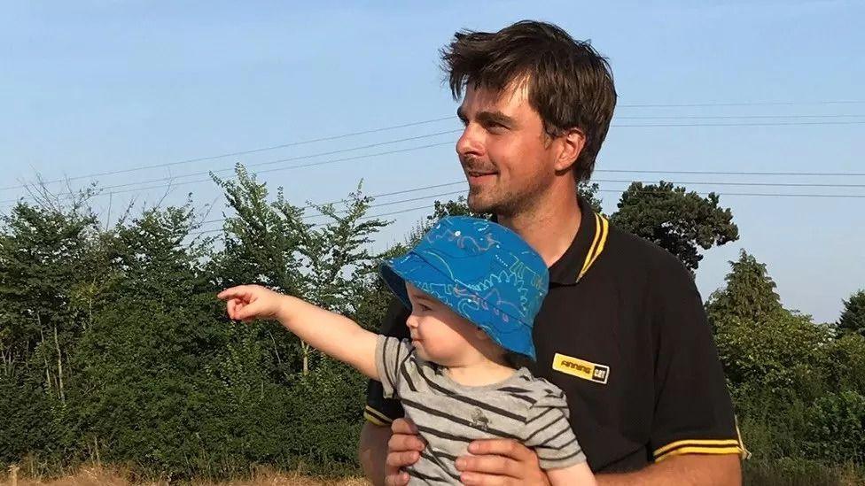 A picture of Jack Last holding his nephew Reggie who is pointing into the distance while the pair are standing in front of trees and bushes