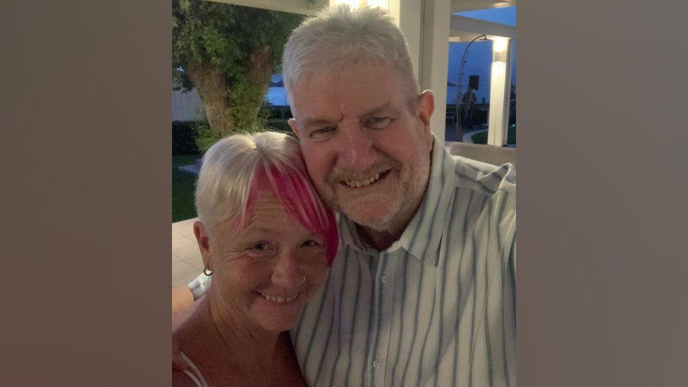 Tim McCarthy is wearing a white shirt with blue and green stripes has a grey beard and thick white hair. Hos wife has short blonde hair with a dyed plum coloured fringe.