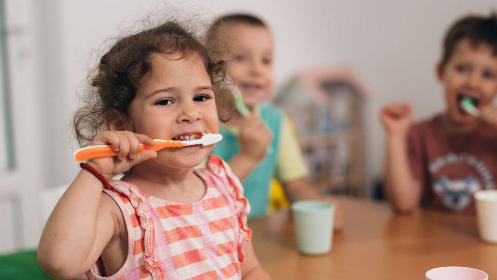 Young children sat around a table brushing teeth. Image focuses on young girl with curly grown hair and striped peach tshirt, smiling as she brushes her teeth. There are pastel coloured plastic beakers on a wooden table and two young boys in the background who are also brushing their teeth