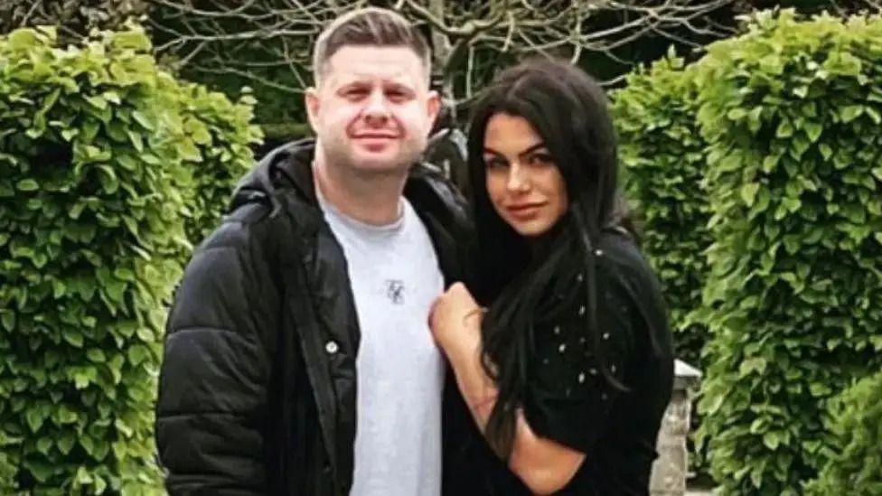 Josh and Chloe Bashford standing in an embrace in front of a hedge. He has short light hair and is wearing a grey t-shirt under a black puffer coat. She has long black hair and is wearing a short-sleeved black jumper