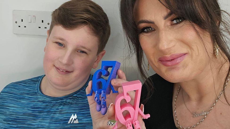 Karlla, a woman with dark hair, andAlfie, a pre-teen boy, sit side-by-side. They are each holding up small plastic statues of the number '67' with little arms and legs attached. The numbers appear in reverse, as in a mirror image, as they have taken a selfie.
