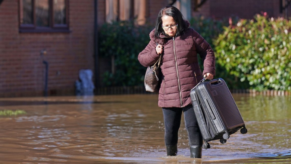 A woman in a purple padded jacket struggles with a black suitcase in flood-water. The water is up to her ankles as she wades through in wellies with a bag over one shoulder. Behind her is the wall of a house and bushes, with water covering the ground around her