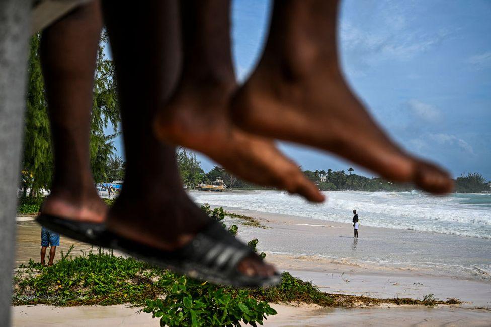 A man wearing white shorts walks on a flooded beach, the legs of people sat on a wall can be seen in the foreground, after the passage of Hurricane Beryl in Bridgetown, Barbados on July 1, 2024