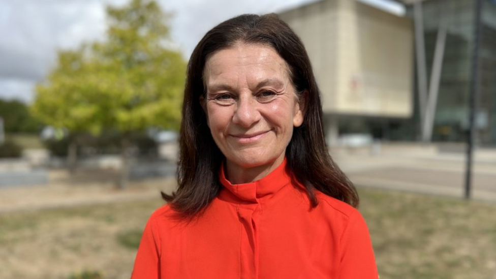 Ms Smith is smiling at the camera from outside the council office building. She wears a bright burnt orange colour top and has shoulder length brown hair. 