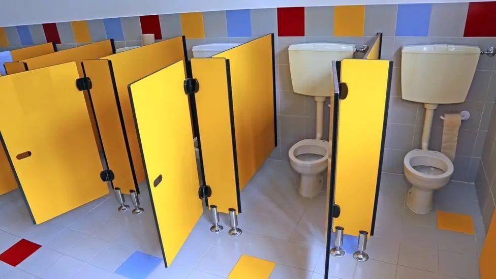 Five toilet cubicles with the doors open with white toilets inside. The doors and walls of the cubicles are yellow. The floor is light blue with some coloured squares on it.