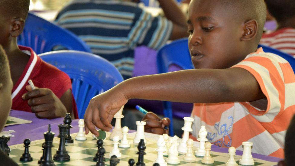 A boy in an orange and white horizontal stripped T-shirt moves a white chess piece on a board in a room where various games are being played.