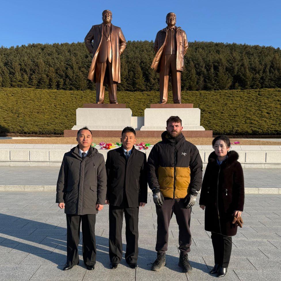 Mike O'Kennedy stands with three North Korean tour guides, with a statue of two men in the background and trees 