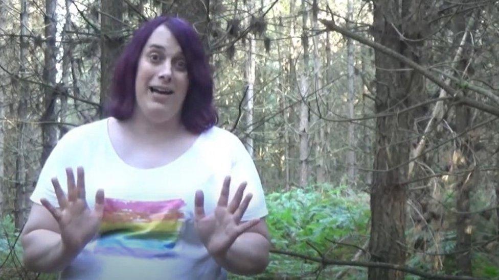 Zoe Watts stands in a forest surrounded by trees. She is wearing a white t-shirt with a rainbow flag on it.