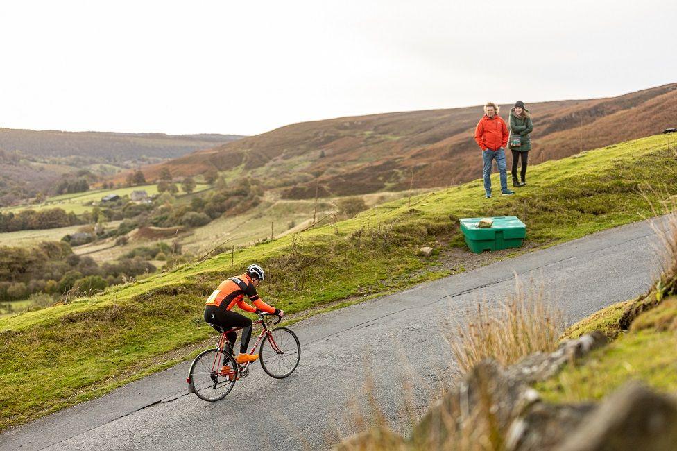 Two people stand by the side of the road as they watch a cyclist battle up the hill