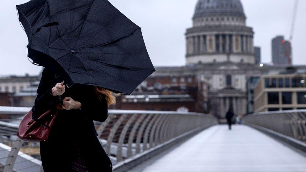 A view of the Millennium Bridge in London, spanning the River Thames under an overcast sky with St. Paul's Cathedral standing prominently in the background. A woman crosses the bridge dressed in black with a black umbrella that has been blown inside out.