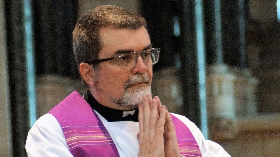 Alan Hodgson standing inside St John the Baptist Roman Catholic Cathedral. He has short silvery dark hair and a silvery goatee beard and is wearing dark-framed glasses, a white surplice with a pink stole over his shoulders and beneath the surplice can be seen a black and white dog-collar. His hands are clasped in prayer and he standing slightly side on